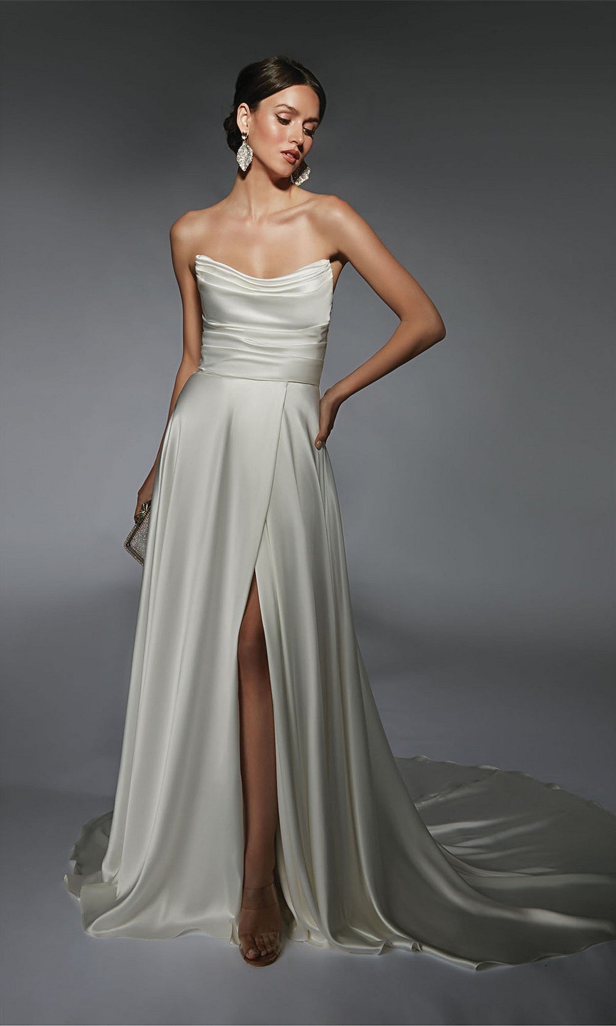 A woman stands against a gray background wearing the Alyce 7107 long white dress with a strapless satin bodice, draping, high slit, and small train. She accessorizes with large earrings and a clutch.
